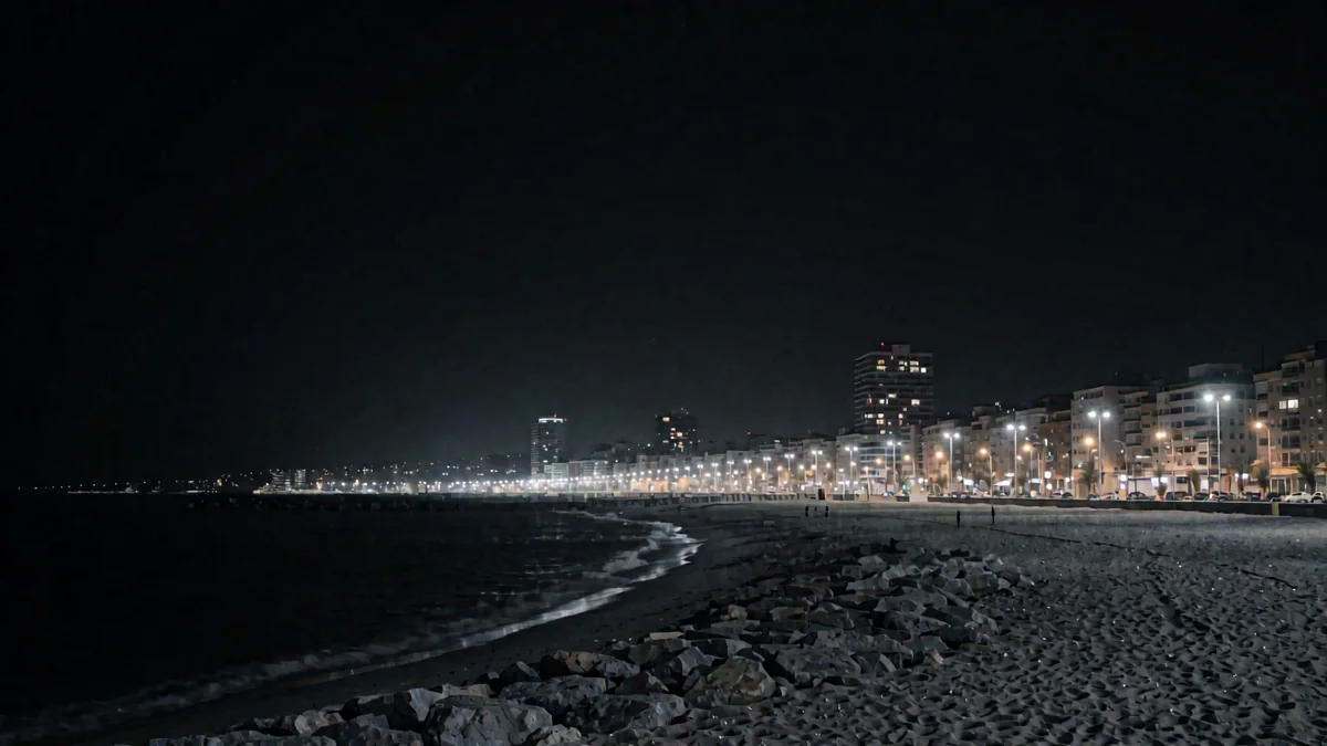 Una playa urbana con un espigón y luces cálidas de una avenida costera en hora azul