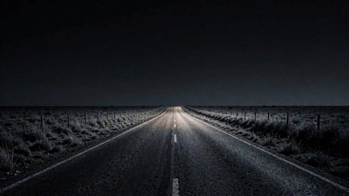 Una ruta recta entre praderas de tambo con alambrados y una luz cálida lejana en hora azul